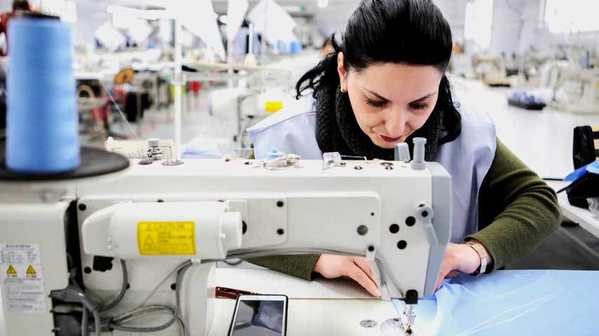Mulher trabalhando em confecção e costurando em máquina reta industrial.