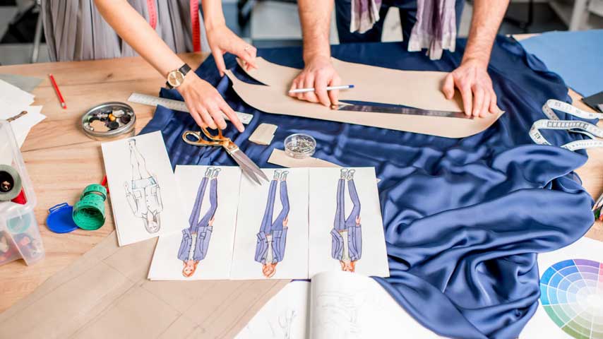 Mesa com croquis, tecido azul, tesoura, fota métrica e duas mãos femininas trabalhando em ateliê na modelagem de roupas. 