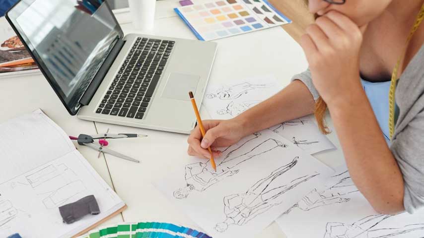 Mulher sentada em frente um notebook e desenhando manualmente croqui de moda.